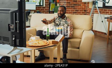 afroamerikanischer Erwachsener, der Bier trinkt und Pommes isst, und eine Comedy-tv-Show im Wohnzimmer genießt. Ein moderner Mann, der Spaß mit einem Film im Fernsehen hat und Fast Food zum Mitnehmen zu Hause serviert. Stockfoto