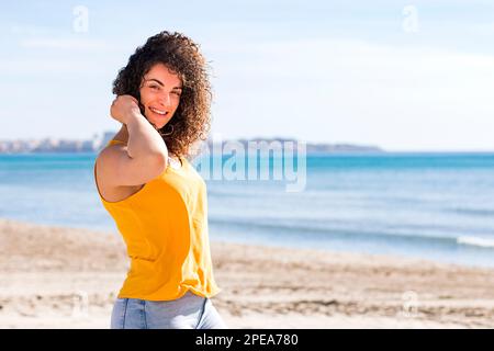 Fröhliche junge, lockige Frau mit gelbem Oberteil, die lächelt und in die Kamera schaut, während sie den Sommerurlaub an der sandigen Küste genießt Stockfoto