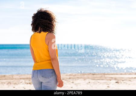 Eine anonyme, lockige Frau mit gelbem Oberteil und Jeans, die am Sandstrand stehen und während der Sommerferien das gewellte Meer bewundern Stockfoto