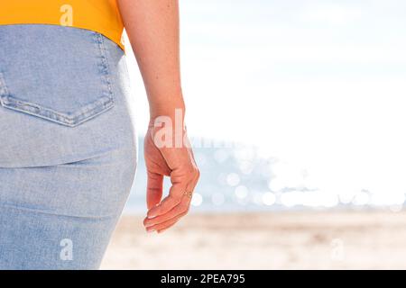 Anonyme Frau in gelbem Hemd und Jeans, die am Sandstrand in der Nähe des Wellenmeers an sonnigen Tagen während der Sommerferien stehen Stockfoto