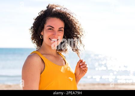 Glückliche Frau lächelt und schaut in die Kamera, während sie im Sommer lockiges Haar auf einem unscharfen Hintergrund des ruhigen Meeres berührt Stockfoto