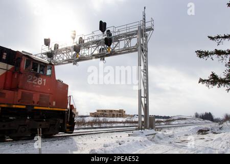 Eine rote Lokomotive unterquert die Bahnlichter, während die Sonne an einem Wintertag durch Wolken scheint, Schnee auf dem Boden. Stockfoto