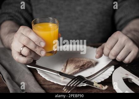 Frühstückszeit. Ein unbekannter Mann, der ein Glas Orangensaft in der Hand hält. Das Sandwich ist auf dem Teller. Stockfoto