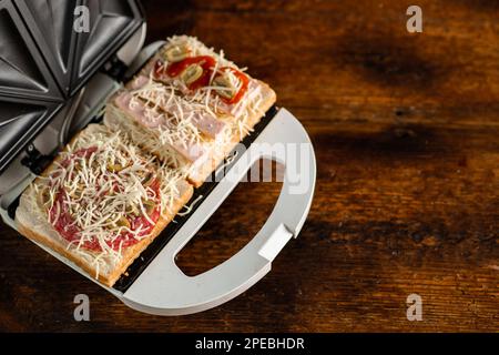 Sandwiches mit Käse, Würstchen und Oliven in einem Sandwichmaker auf Holzhintergrund zubereiten. Stockfoto