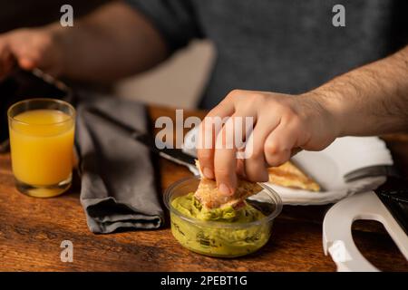 Ein unbekannter Mann isst Toast mit Guacamole. Frühstückszeit. Die Hände des Mannes halten einen Toast. Stockfoto