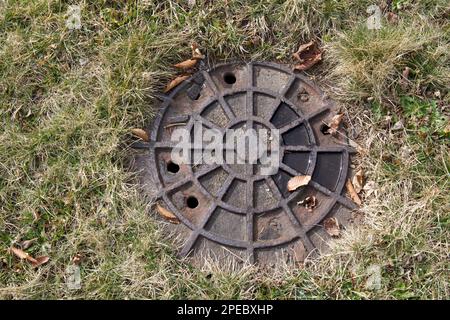 Alte Kanalisation oder Verschlusskappe, verrostet. Im Winter in New England im Gras mit getrockneten Blättern sitzen. Stockfoto