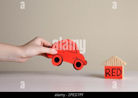 Eine Frau, die ein rotes Auto in der Nähe eines Holzhauses hält, am weißen Tisch, Nahaufnahme. ABA-Therapiekonzept Stockfoto