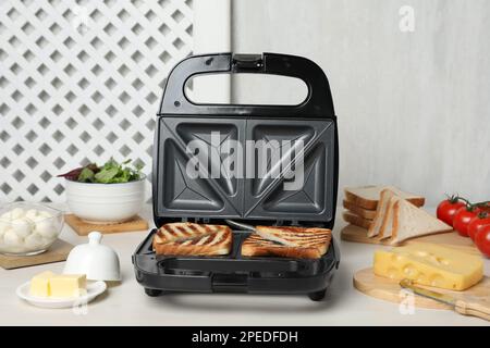 Moderne Grillmaschine mit Sandwiches und verschiedenen Produkten auf einem weißen Tisch Stockfoto