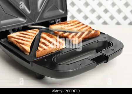 Moderner Sandwichmaker mit Brotscheiben auf weißem Holztisch Stockfoto