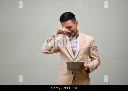 Trauriger und weinender asiatischer Geschäftsmann oder männlicher CEO in formellen Business-Anzügen steht vor einem grünen Studiohintergrund mit seinem Tablet, das sich wischte Stockfoto