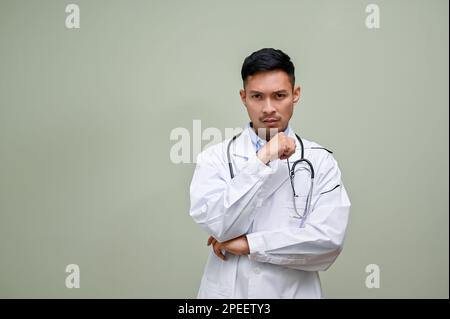 Gestresster und aufmerksamer asiatischer Millenialarzt in einem weißen Kleid, der seine Brille in der Hand hält, den Arm gekreuzt und dabei mit einem ernsten Gesicht in die Kamera schaut Stockfoto