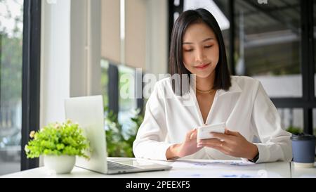 Attraktive asiatische Millennials-Geschäftsfrau oder Buchhalterin, die mit einem Taschenrechner Umsätze berechnet und an ihrem Schreibtisch im Büro arbeitet. Stockfoto