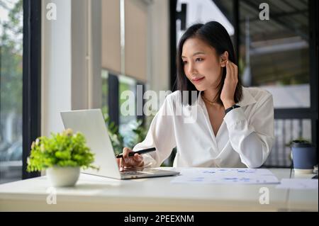 Eine charmante und erfolgreiche asiatische Geschäftsfrau, die im Büro an ihrem Unternehmensprojekt mit einem Notebook arbeitet. Stockfoto