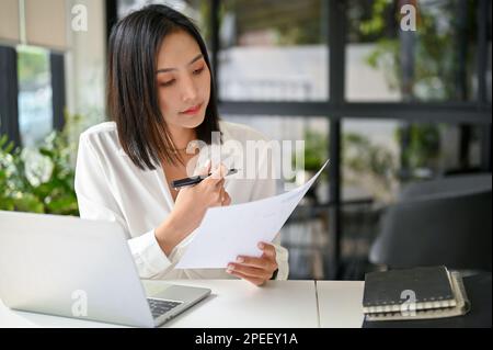 Fokussierte und professionelle asiatische Geschäftsfrau oder Chefin der Millennials, die Geschäftsdokumente prüft oder Finanzdatenberichte analysiert und bei ihr arbeitet Stockfoto