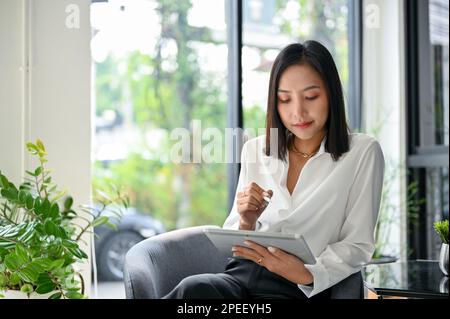 Charmante asiatische Geschäftsfrau oder Chefin der Millennials, die ihr digitales Tablet benutzt, während sie sich im Café entspannen. Stockfoto