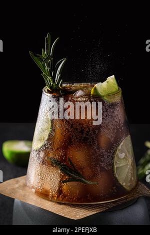 cola mit Limette und Eis auf dunklem Hintergrund. Dekoriert mit einem Zweig Rosmarin und einer Scheibe Limette Stockfoto