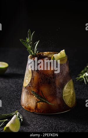 cola mit Limette und Eis auf dunklem Hintergrund. Dekoriert mit einem Zweig Rosmarin und einer Scheibe Limette Stockfoto