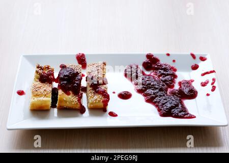 Auf einem weißen rechteckigen Teller werden ein Kuchen und ein mit Marmelade bestreuter Kuchen ungenau mit roten Marmeladensamen bestreut. Das Essen wird schlecht serviert, so ein Eindruck wie Blut Stockfoto