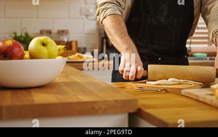 Nahhände Mann mit Schürze, der einen Rollnadel hält und Teig auf dem Küchentisch macht. Stockfoto