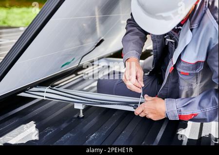 Nahaufnahme eines männlichen Arbeiters, der Kabel mit einem speziellen Kabelbinder sichert. Installation von Solarbatterien auf dem Dach des Hauses. Ein Ingenieur in einem weißen Helm, der alle Kabel des Solar-PV-Panels zusammenhält. Stockfoto