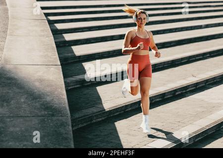 Sportlerin, die draußen mit Kopfhörern laufen will. Läuferin, die in Sportbekleidung die Treppe runterjoggt. Weibliches Training mit Herz-Kreislauf-Übungen. Stockfoto