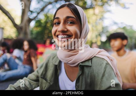 Fröhliches muslimisches Mädchen, das vor der Kamera lächelt, während es bei einem Klimaprotest mit einer Gruppe von Demonstranten saß. Multikulturelle Jugendaktivisten schließen sich der an Stockfoto