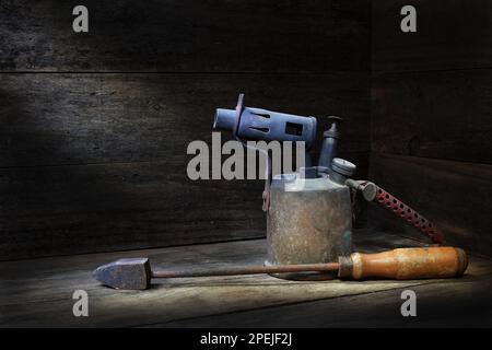 Ein klassischer Lötkolben aus Messing, handgepumpt, Kerosin/Gas-Lötlampe und Lötkolben mit Kupferspitze in rustikalem Holz in sanfter, dunkler Stimmungsbeleuchtung Stockfoto