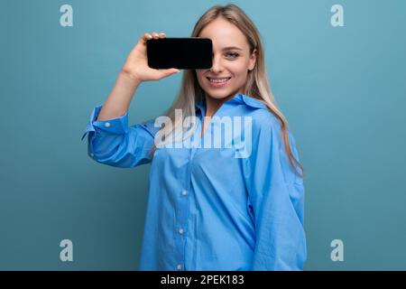 Lächelndes blondes Mädchen, das ein Smartphone mit leerem Bildschirm auf blauem Hintergrund hält Stockfoto