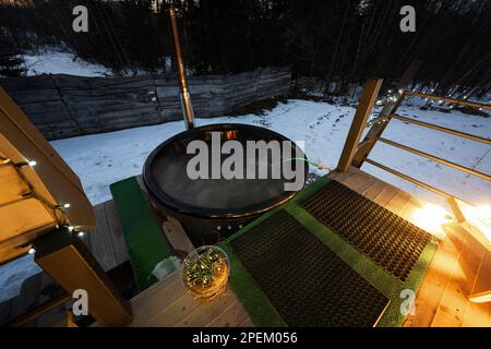 Wannen-Whirlpool mit Holzfass auf der Terrasse der Hütte in der Winternacht. Skandinavische Badewanne mit Kamin zum Brennen von Holz und zum Heizen von Wasser. Stockfoto