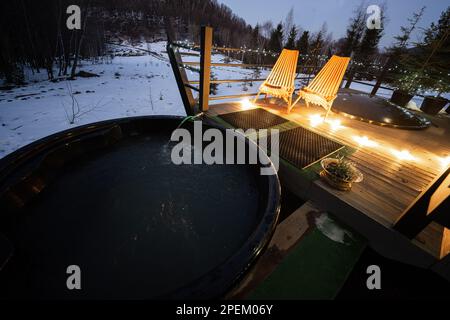 Wannen-Whirlpool mit Holzfass auf der Terrasse der Hütte in der Winternacht. Skandinavische Badewanne mit Kamin zum Brennen von Holz und zum Heizen von Wasser. Stockfoto