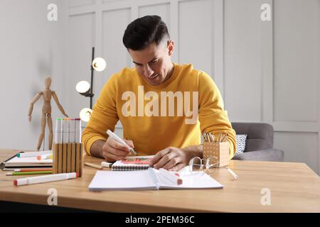 Ein Mann, der im Skizzenbuch mit Filzstift an einem Holztisch im Innenbereich malt Stockfoto