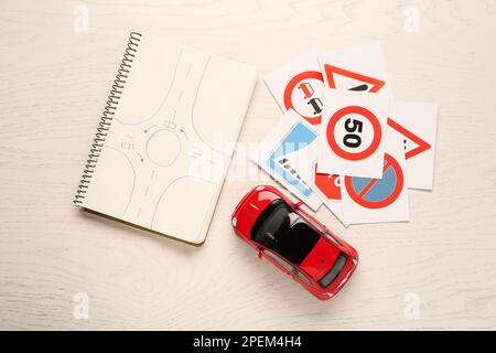 Viele verschiedene Straßenschilderkarten, Notizbuch mit Skizze des Kreisverkehrs und Spielzeugauto auf weißem Holztisch, flach liegend. Fahrschule Stockfoto