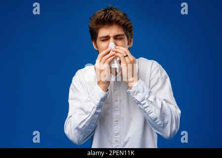 Ein junger Mann niest in eine Serviette auf blauem Hintergrund Stockfoto
