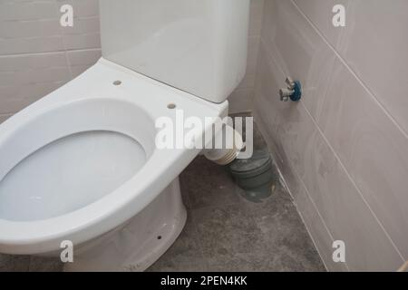 Nahaufnahme der weißen Toilettenschüssel und der Leitungen, die den Anschluss der Toilettenpfanne mit dem Abflussrohr in der Ecke des Badezimmers verbinden. Stockfoto