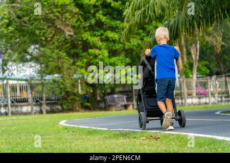Ein kleiner Junge schubst ein Baby in einem Kinderwagen. Das Kind lernt, sich um den jüngeren Bruder oder die jüngere Schwester in der Familie zu kümmern und Verantwortung dafür zu übernehmen. In Stockfoto