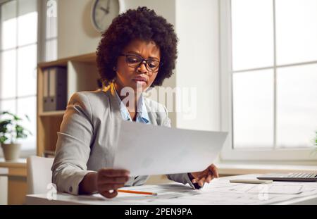 Eine seriöse Geschäftsfrau, die am Schreibtisch sitzt und sich Papierdokumente ansieht Stockfoto
