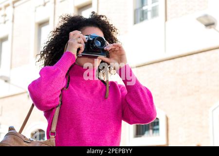 Kleiner Winkel einer jungen kaukasischen Frau in pinkfarbenem Rollkragenpullover mit Tasche, die an sonnigen Tagen auf der Straße der Stadt steht und Fotos mit einer Filmkamera macht Stockfoto