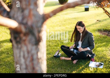 Lächelnde, attraktive Geschäftsfrau der Millennials, die auf grünem Gras im Park sitzt, Online-Schulung, Remote-Arbeit auf einem Laptop. Kaffeepause, Ruhe und Entspannung Stockfoto