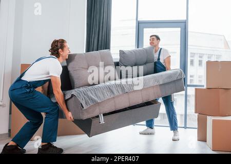 Schweres Sofa. Zwei junge Spielfiguren in blauer Uniform, die im Zimmer arbeiten Stockfoto