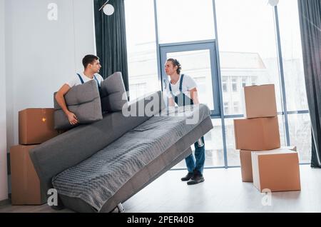 Schweres Sofa. Zwei junge Spielfiguren in blauer Uniform, die im Zimmer arbeiten Stockfoto