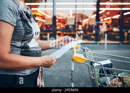 Frau, die nach dem Einkaufen im Supermarkt auf der Quittung nach höheren Preisen sucht. Eine Frau steht vor dem Markt mit einem Einkaufswagen und prüft ihn sorgfältig Stockfoto