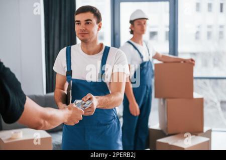 Der Mann nimmt Geld von einem anderen Kerl. Zwei junge Spielfiguren in blauer Uniform, die im Zimmer arbeiten Stockfoto