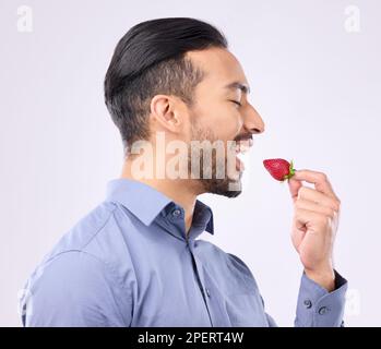 Gesunde Ernährung und ein Mann, der eine Erdbeere im Studio auf grauem Hintergrund für natürliche Ernährung isst. Essen, Obst und Veganer mit einem gutaussehenden jungen Mann Stockfoto