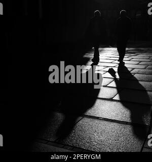 Eine Graustufenaufnahme von Menschen, die in der Dämmerung die Straße entlang gehen. Stockfoto