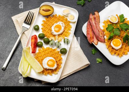 Zwei Teller mit Frühstück. Waffeln, Speck, Oliven und Wachteleier. Flach verlegt. Schwarzer Hintergrund. Stockfoto