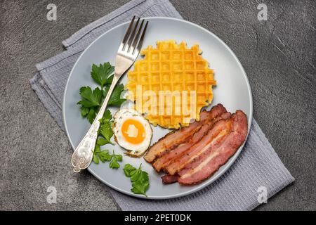 Gebratener Speck mit Ei, Waffeln und Petersilie-Zweigen auf einem grauen Teller. Graue Serviette. Flach verlegt. Schwarzer Hintergrund. Stockfoto