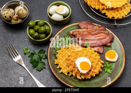 Wachteleier und grüne Oliven in Schüsseln. Gebratener Speck, Waffeln und Eier in einer Bratpfanne. Schwarzer Hintergrund Stockfoto