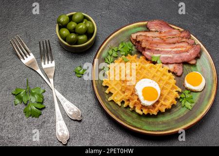 Grüne Oliven in einer grünen Schüssel. Zwei Gabeln auf dem Tisch. Gebratener Speck, Waffeln und Eier auf einem grünen Teller. Stockfoto