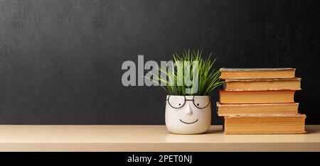 Ein Stapel alter Bücher und eine Topfpflanze auf dem Tisch vor der Wand der Tafel. Mit Platz für Ihren Text. Lern- und Arbeitskonzept Stockfoto
