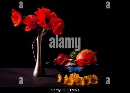 Stillleben auf schwarzem Hintergrund. Ein Strauß wilder roter Mohnblumen in einem Leuchtfeuer. Schale mit Früchten - Granatäpfel, Apfel und Avocado. Speicherplatz kopieren. Stockfoto